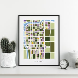May include: A framed art print of a colorful, abstract map labeled "THE LOOP." The map features a grid-like design with blocks of green, purple, yellow, and blue. A cactus in a white pot and a black clock are on a white surface.