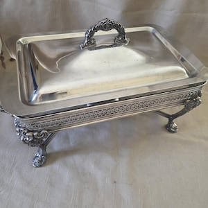 Puede incluir: Una fuente rectangular plateada con tapa y asa ornamentada. La fuente se apoya en cuatro patas decorativas. La tapa y la base tienen patrones detallados. Adecuada para servir alimentos en un evento formal.