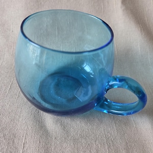 May include: A clear, round, blue glass teacup with a handle. The cup has a rounded body and a small handle. The glass is a vibrant blue color. The cup is sitting on a light-colored, textured surface.