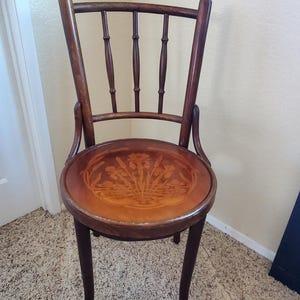 May include: A vintage wooden chair with a round seat featuring a floral design. The chair has a dark brown finish, curved back, and four slender legs. The seat is a lighter brown color.
