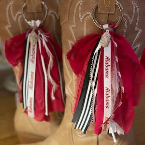 May include: A pair of tan cowboy boots with decorative stitching. Each boot has a silver ring attached to a tassel featuring red fabric, ribbons with "Alabama" text, black and white striped ribbon, and a sparkling charm.