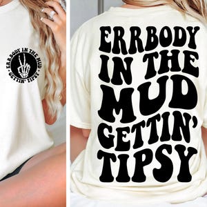 以下が含まれることがあります： 背面に黒の太字レトロフォントで「ERRBODY IN THE MUD GETTIN' TIPSY」と書かれたクリーム色のTシャツ。前面には、骸骨の手が平和のサインを作っている円形のグラフィックがあります。カジュアルで楽しいTシャツ。