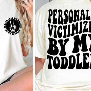 Peut inclure: T-shirt blanc avec un texte noir qui dit "PERSONALLY VICTIMIZED BY MY TODDLER" dans une police rétro. Le texte est centré sur le t-shirt.