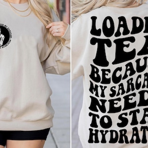 May include: A beige sweatshirt with black text that reads "Loaded Tea Because My Sarcasm Needs To Stay Hydrated". The text is in a retro font style. The front of the sweatshirt has a smaller black graphic of a skeleton hand making a peace sign.