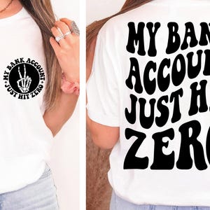Puede incluir: Camiseta blanca con un signo de paz negro y el texto "My Bank Account Just Hit Zero" en la parte delantera. La parte posterior de la camiseta tiene el texto "My Bank Account Just Hit Zero" en una fuente retro.