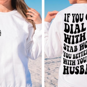 May include: White sweatshirt with a black graphic on the front that says "Flirt with your own husband" and a peace sign with a skeleton hand. The back of the sweatshirt has a black graphic that says "If you can't dial 911 with six stab wounds you better flirt with your own husband."