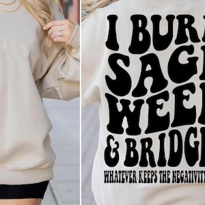 May include: A cream-coloured sweatshirt with black text and graphics. The front has a circular design with a skeleton hand giving a peace sign and the words "Whatever Keeps The Negativity Away". The back says "I Burn Sage Weed & Bridges" in large, bold letters.