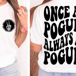 Puede incluir: Camiseta blanca con un signo de paz negro con una mano de esqueleto y el texto "Once a Pogue Always a Pogue" en la parte delantera. La parte posterior de la camiseta tiene el texto "Once a Pogue Always a Pogue" en una fuente retro.