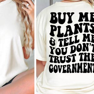 May include: A white t-shirt with black text that reads "Buy me plants & tell me you don't trust the government".
