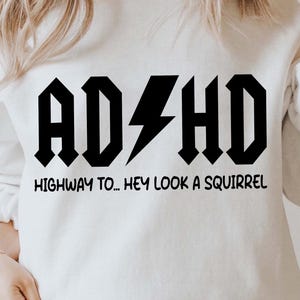 Op de afbeelding: Wit sweatshirt met zwarte tekst die "ADHD HIGHWAY TO... HEY LOOK A SQUIRREL" luidt. De tekst is in een vetgedrukt, rock-'n-roll-lettertype.