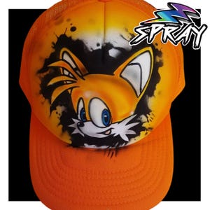 May include: Orange and black trucker hat with a spray-painted image of a cartoon fox with a white and blue face. The text "SPRAY" is in the upper right corner of the hat.
