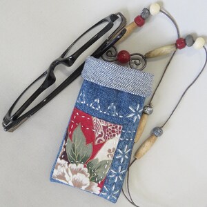 May include: A denim eyeglasses case with a floral patchwork design and a beaded cord. The case is blue with white stitching and a gray patterned top. Black eyeglasses rest beside the case.