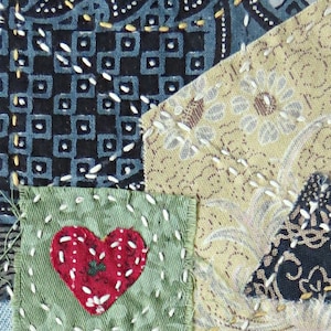 May include: A patchwork quilt block with a green fabric square in the center featuring a red heart. The heart is surrounded by white stitching. The green square is surrounded by other fabric squares with different patterns and colors, including blue, beige, and black.