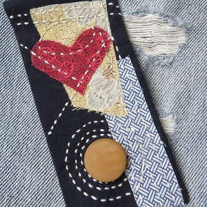 May include: A dark blue fabric patch with a red heart, white stitching, and a brown button. The patch is sewn onto a light blue denim fabric.