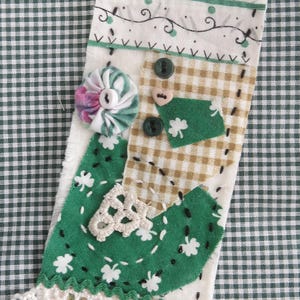May include: A handmade fabric ornament with a green, white, and brown patchwork design. The ornament is decorated with a white crocheted trim, a green shamrock, and the word "Irish" written in black.
