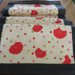 May include: A white table runner with a black border and a floral pattern of red and white flowers. The flowers are stylized and have a retro look.