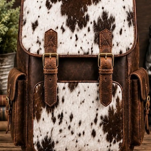 Puede incluir: Una mochila de cuero marrón con un patrón de piel de vaca blanca y marrón en la solapa superior y el bolsillo frontal. La bolsa incluye dos bolsillos laterales, un asa superior y dos correas de cuero verticales con hebillas de metal bronce.