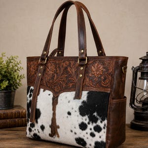 May include: A structured tote bag featuring a combination of dark brown leather with intricate floral tooling on the upper section and a black and white cowhide panel on the lower section, complete with two leather handles and side pockets.