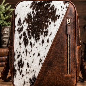 May include: A backpack featuring a front panel of brown and white cowhide hair-on leather contrasted with distressed brown leather. The design includes a vertical zippered pocket on the right side, a top handle, and gold-toned metal hardware.