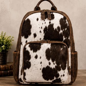 Può includere: Uno zaino con un motivo a pelle di mucca bianco e marrone scuro sul pannello frontale, completato da finiture in pelle marrone invecchiata, una maniglia superiore, due tasche laterali e due scomparti con cerniera e dettagli in ottone.