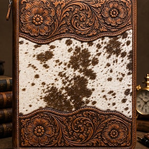 May include: A rectangular leather portfolio featuring a central panel of white and brown cowhide hair-on leather. The top and bottom borders are made of dark brown leather with intricate, embossed floral and scrolling vine patterns.