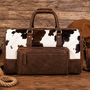 May include: A duffel bag featuring a white and dark brown cowhide pattern on the upper section, with a solid dark brown leather lower panel and front zippered pocket. The bag includes two top handles, brass-toned hardware, and a detachable strap.
