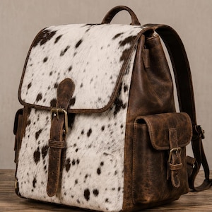 Peut inclure: Un sac à dos doté d'un panneau avant en peau de vache blanche et brun foncé avec une finition texturée. Les côtés, les bretelles et la poignée supérieure sont en cuir brun foncé lisse. Il comprend des boucles en métal doré et des poches latérales.