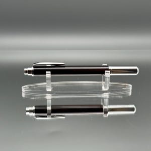 May include: A black and silver pen on a clear acrylic stand. The pen features a dark brown barrel and silver clip and tip. The pen's reflection is visible on the glossy surface beneath it.