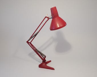May include: Red adjustable desk lamp with a conical shade and articulated arm. The lamp has a rectangular base and a spring-loaded mechanism for adjusting the height and angle. The lamp is made of metal and has a glossy finish.