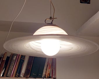 May include: A white glass pendant light with a swirling pattern, featuring a wide, flat, saucer-shaped shade and a central spherical bulb. The fixture is suspended by a thin cord and includes a metallic top attachment with a small loop.