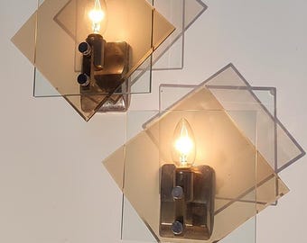 May include: A pair of wall sconces featuring square amber-tinted glass panels mounted at an angle. Each fixture has a central metallic base with a single candle-style light bulb and two small circular knobs on the front of the light housing.