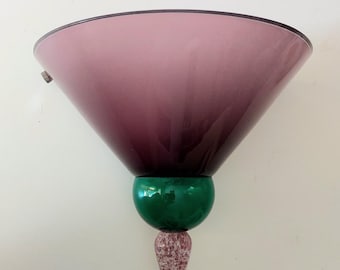 May include: A wall sconce featuring a wide, conical, translucent purple glass shade. The fixture includes a spherical, solid green glass accent piece positioned below the shade, terminating in a pointed, speckled, light-colored glass finial.