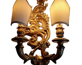 May include: Ornate gold-colored wall sconce with two lights. The sconce features a detailed, sculpted design with a mythical creature's head at the center. Each light has a beige lampshade. A decorative lighting fixture for a home.