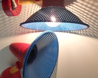 May include: Two wall-mounted lamps featuring red circular bases with black rocker switches, yellow cylindrical arms, and conical shades made of blue perforated metal mesh. The top lamp is illuminated, casting a patterned shadow on the surface.