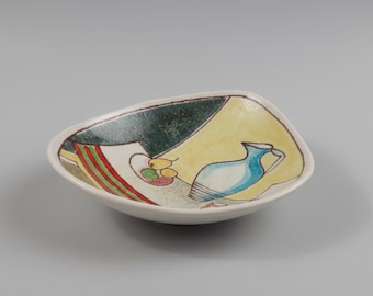 May include: A small, white, ceramic dish with a unique, asymmetrical shape. The dish features a hand-painted design with a yellow background, a blue pitcher, and a still life of fruit. The rim is outlined in a dark brown color.