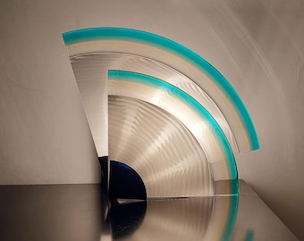May include: Abstract metal sculpture with a fan-like design. The sculpture features curved, brushed metal elements with a teal and white accent along the edges. The piece is displayed on a reflective black surface.