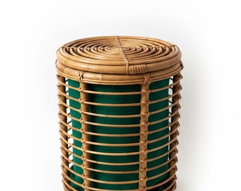 May include: A cylindrical rattan basket featuring a natural light brown finish with a woven lid and vertical slats. The interior is lined with a solid dark green fabric, creating a contrast between the organic wood texture and the deep color.