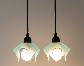 May include: Two pendant lights featuring translucent, pale mint green glass shades with a soft, ruffled, flower-like shape. Each shade is suspended by a black cord and a black socket, housing a single glowing white light bulb in the center.