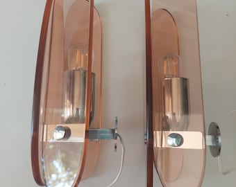 May include: A pair of wall sconces featuring elongated, translucent peach-colored acrylic panels. Each fixture includes a central metallic socket housing a clear light bulb, supported by a horizontal metal arm and a circular wall-mounting plate.