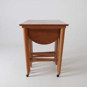 May include: A set of nesting tables made of light brown wood. The tables have rounded legs and a half-circle apron. The tables are on small black wheels. The tables are a mid-century modern style.