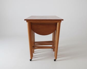 May include: A set of nesting tables made of light brown wood. The tables have rounded legs and a half-circle apron. The tables are on small black wheels. The tables are a mid-century modern style.