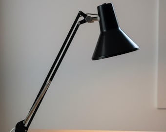 May include: A black architect-style desk lamp with a conical shade and an adjustable articulated arm. The lamp features a clamp base for attachment to a surface and includes a silver-colored spring mechanism along the lower arm section.