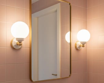 May include: A rectangular wall mirror with rounded corners and a thin gold frame, flanked by two spherical white glass wall sconces with gold accents. Below the mirror is a gold wall-mounted faucet fixture set against light pink square tiles.