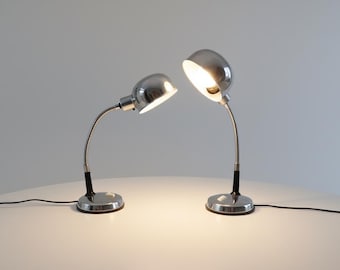 May include: Two chrome desk lamps with flexible gooseneck arms and round bases. The lamps have a polished finish and are positioned on a white surface. One lamp is illuminated, casting a warm glow.