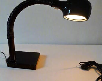 May include: A black desk lamp with a flexible, ribbed neck and a rectangular base. The lamp head is oval-shaped and emits a warm, bright light. A black power cord extends from the base.