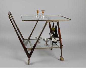 May include: A vintage bar cart with a glass top and lower shelf, supported by a brass-toned frame. The cart features dark wood accents and is equipped with wheels. Two wine bottles and two glasses are visible.