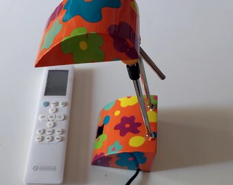 May include: A desk lamp with an orange base and shade featuring a multi-colored floral pattern in blue, green, and purple. The lamp has a chrome-finished adjustable arm and a black power cord, positioned next to a white Olimpia Splendid remote.