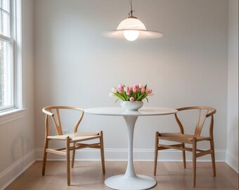 May include: A dining room set featuring a white tulip table, two wooden chairs with woven seats, and a floral arrangement. A white and glass pendant light hangs above the table. The room has light-colored walls and a window.