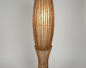 May include: A tall floor lamp featuring a cylindrical bamboo cage structure with vertical slats. The lamp has a flared base and a wider upper section, enclosing a soft, glowing white inner light diffuser that illuminates the bamboo frame.