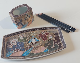 May include: An ornate, rectangular ceramic tray and a small, square ceramic container with a pencil holder, featuring detailed illustrations of figures in traditional attire. The tray and container are decorated with gold accents and a dark brown border. Four black pencils are also present.